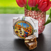 Panforte Cake in hand wrapped waxed paper. Slices of cake showing chunks of citrus peel and creamy white nuts. Flower design glass vase holding red tulips.