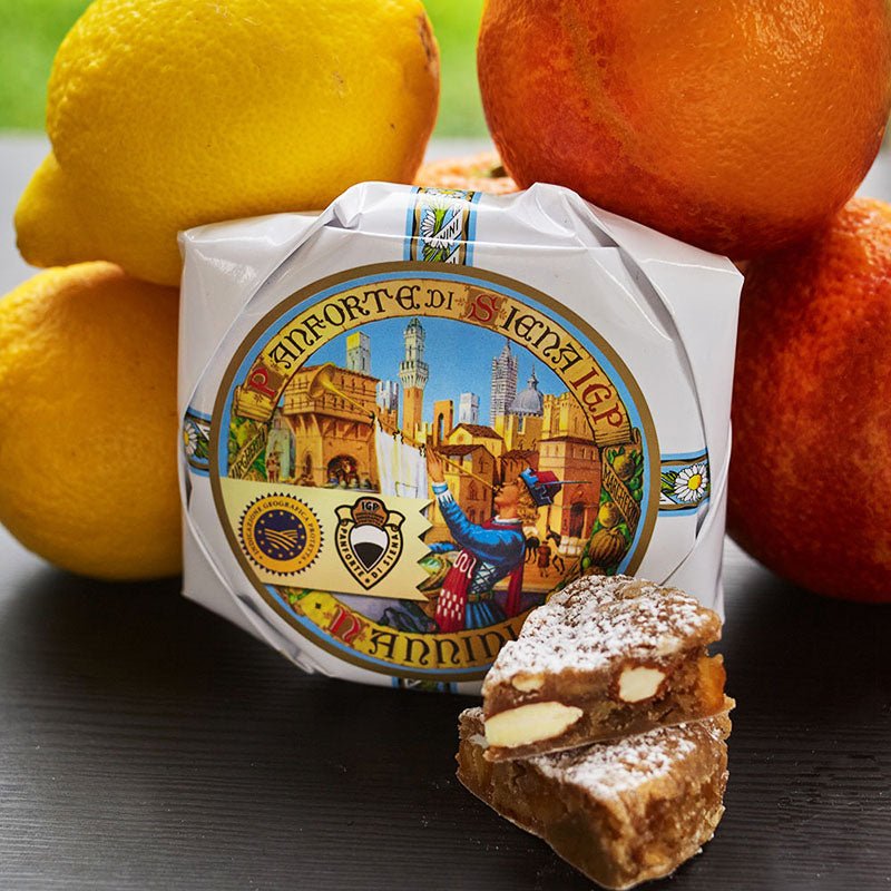 Panforte Cake in hand wrapped waxed paper. Slices of cake showing chunks of citrus peel and creamy white nuts. With Oranges and Lemons.